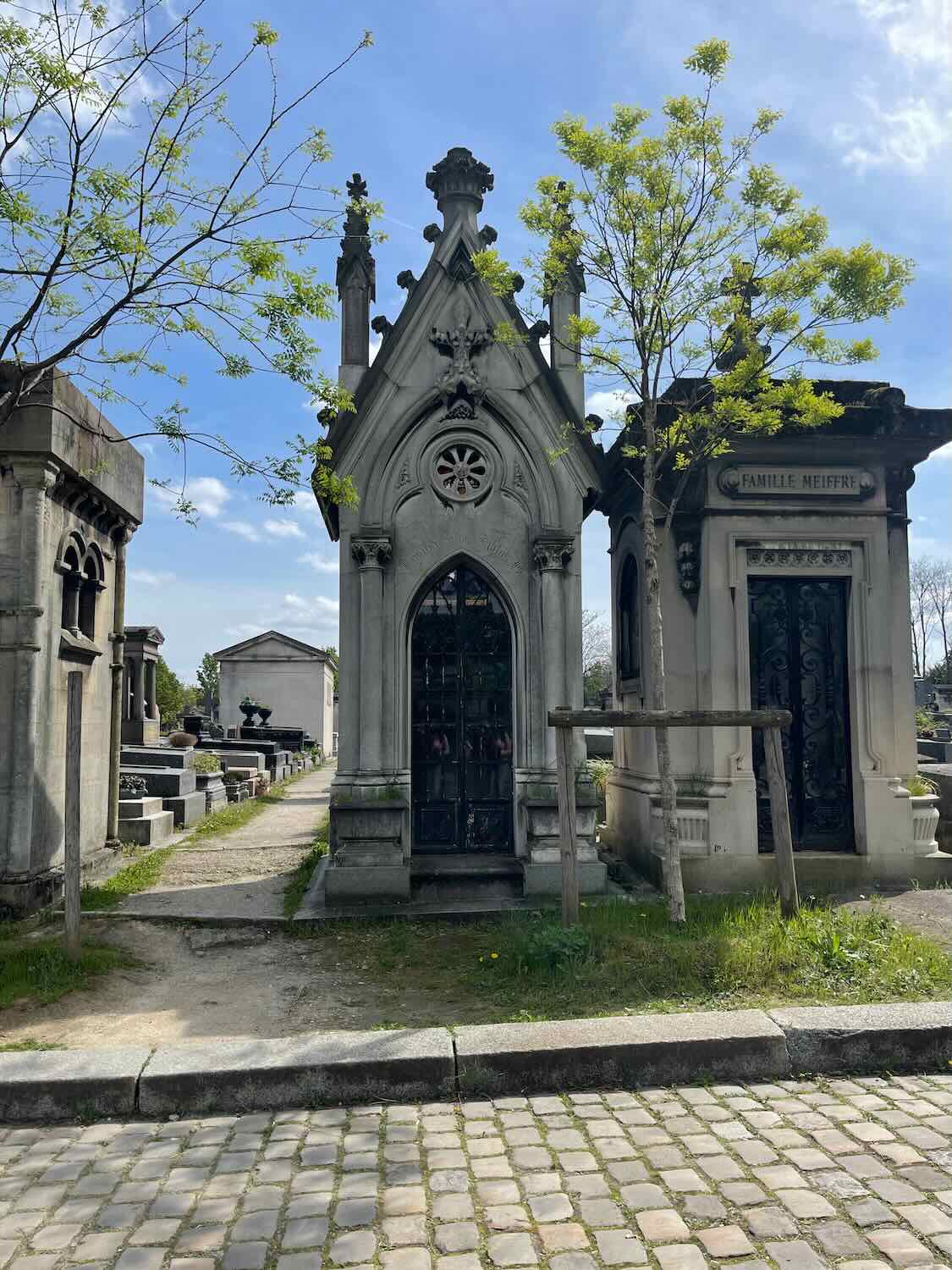 A serene pathway through a historic Parisian cemetery, featuring ornate family mausoleums and vibrant green trees, reflecting the peaceful and solemn atmosphere of the site.