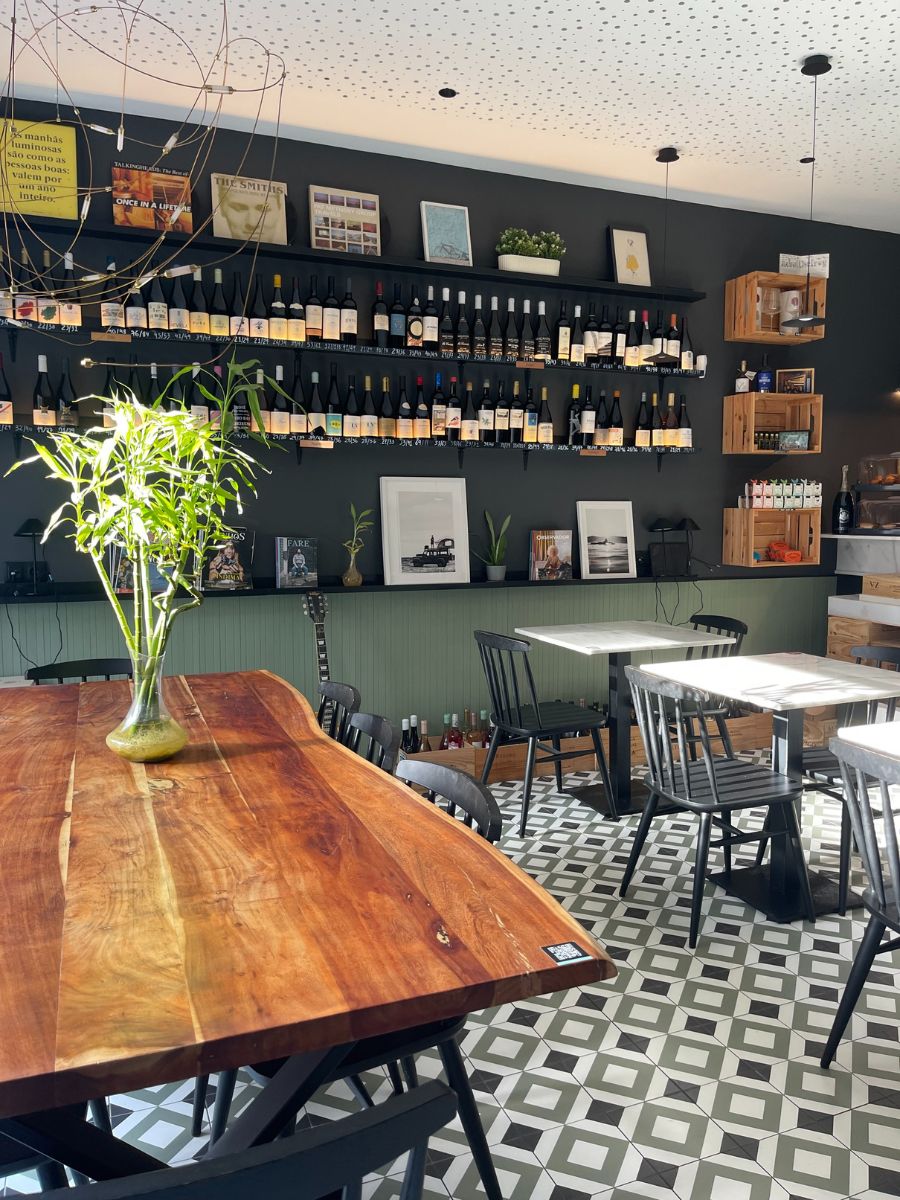 The interior of a bright wine bar, featuring a long live-edge wooden communal table topped with a vase of bamboo, black dining chairs on a patterned tile floor, and dark walls lined with shelves of assorted wine bottles and framed prints.