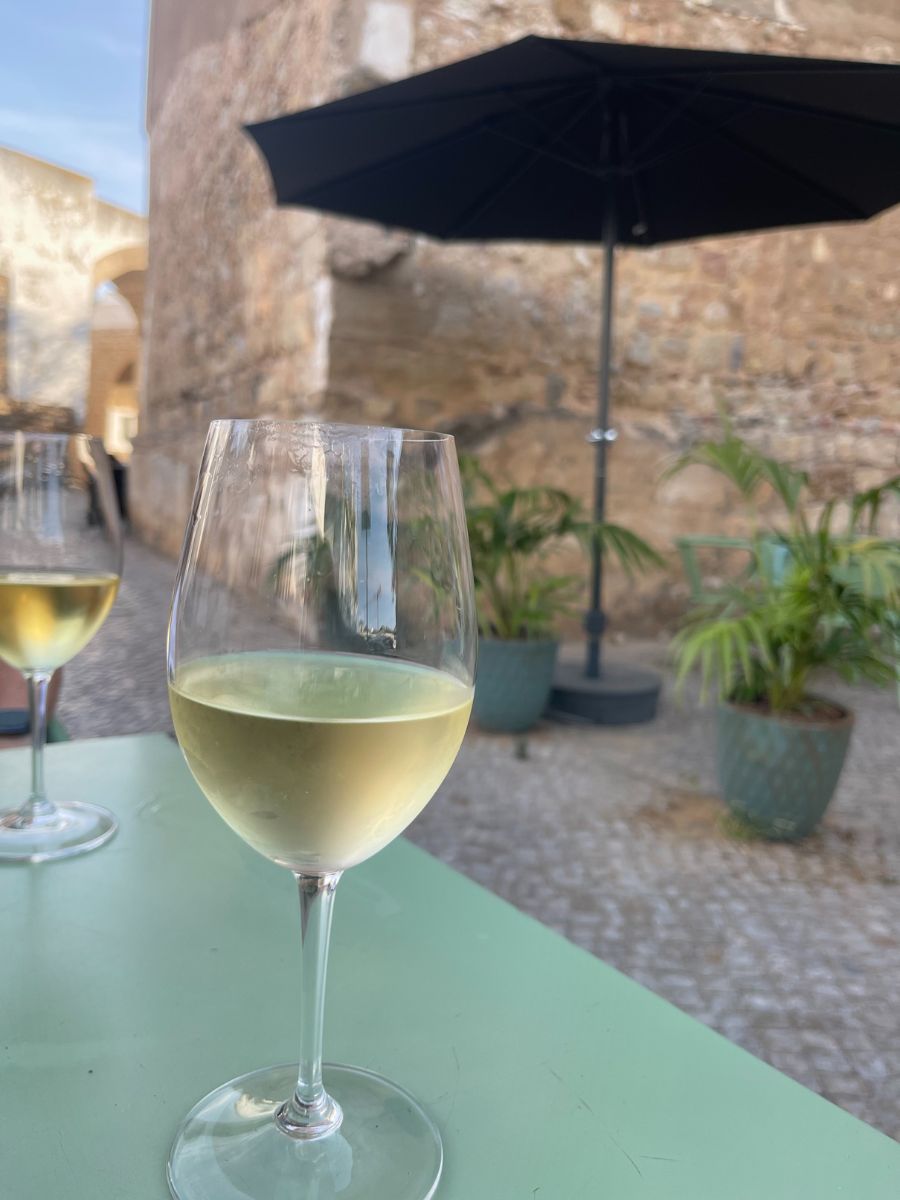 A close-up of a half-full glass of chilled white wine sitting on a pale green café table, with a second wine glass, potted plants, a black patio umbrella, and a weathered stone wall in soft focus behind it.
