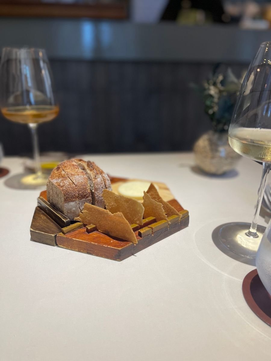 A wooden board display at a white-clothed table holding a sliced round loaf of rustic bread, triangular crisp wafers, and a dish of whipped butter, flanked by stemmed wine glasses and a vase of dried foliage.