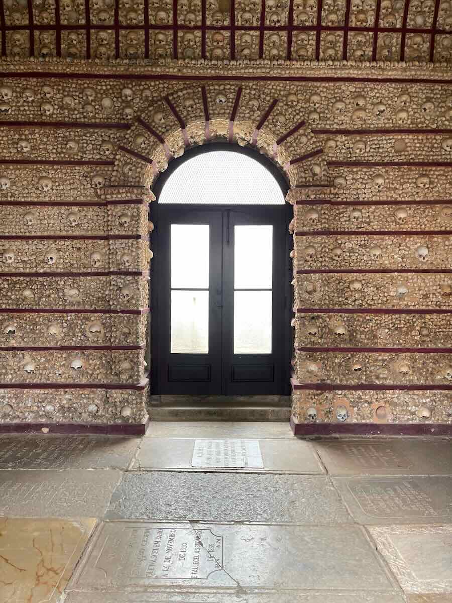 A somber interior in Faro, featuring a wall intricately decorated with human skulls and bones, surrounding an arched doorway with glass panels, conveying a historical and contemplative atmosphere.