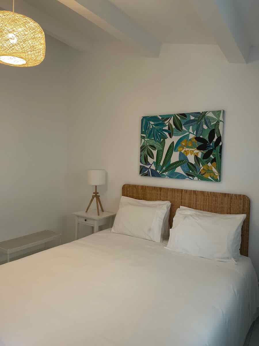 A cozy bedroom in Faro with a wicker headboard, white bedding, and a bedside table holding a tripod lamp. A colorful painting of tropical leaves hangs above the bed, and a woven ceiling light adds warmth to the space.