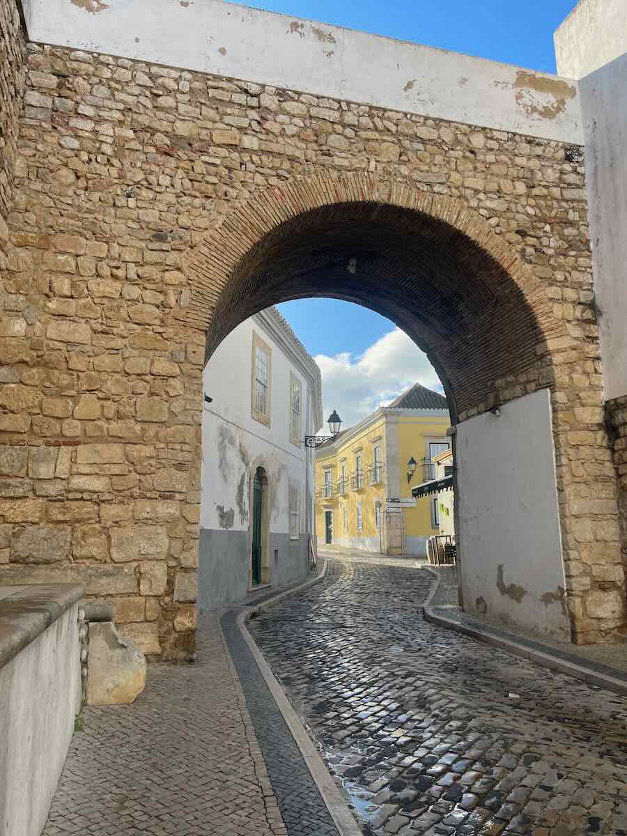 A cobblestone street passing under a rustic stone archway, leading to pastel-colored buildings with traditional architecture, bathed in sunlight in Faro, Portugal.