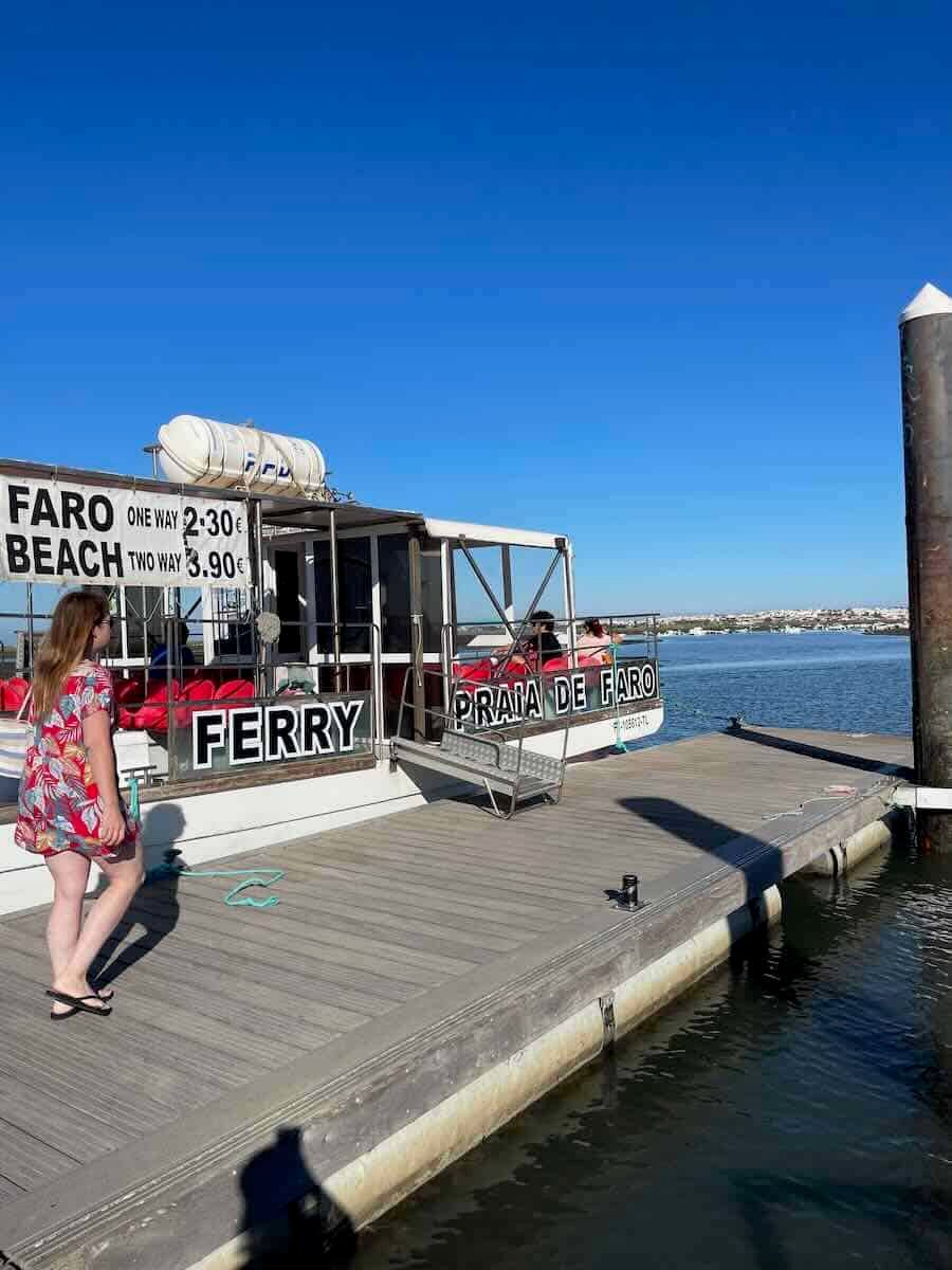 Docked ferry labeled 'Praia de Faro' with ticket prices displayed, a person walking nearby, and calm water under a clear blue sky.