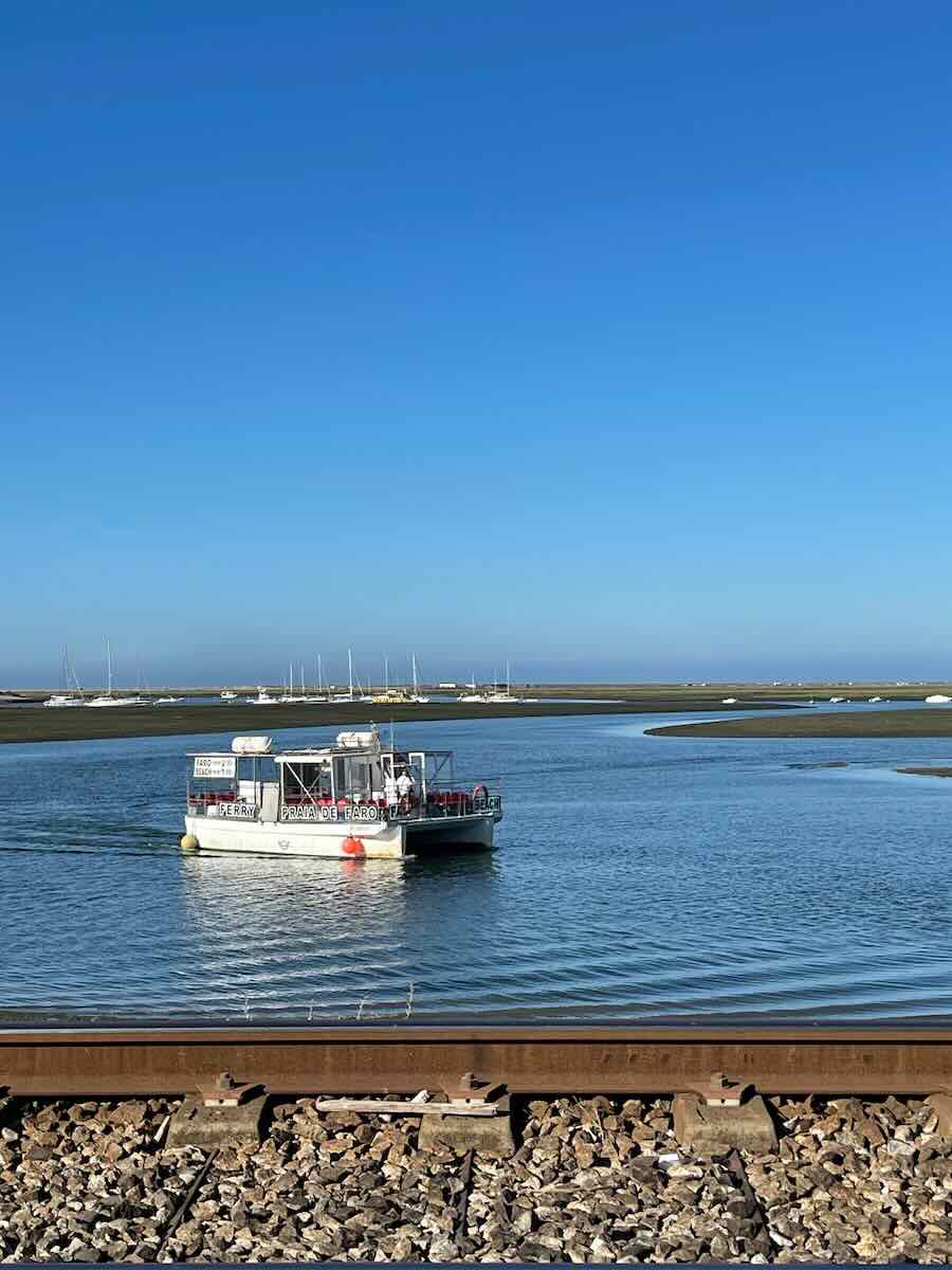 A small ferry boat labeled "Praia de Faro" navigates through calm blue waters, with moored boats and sandbanks in the background. A railway track is visible in the foreground.