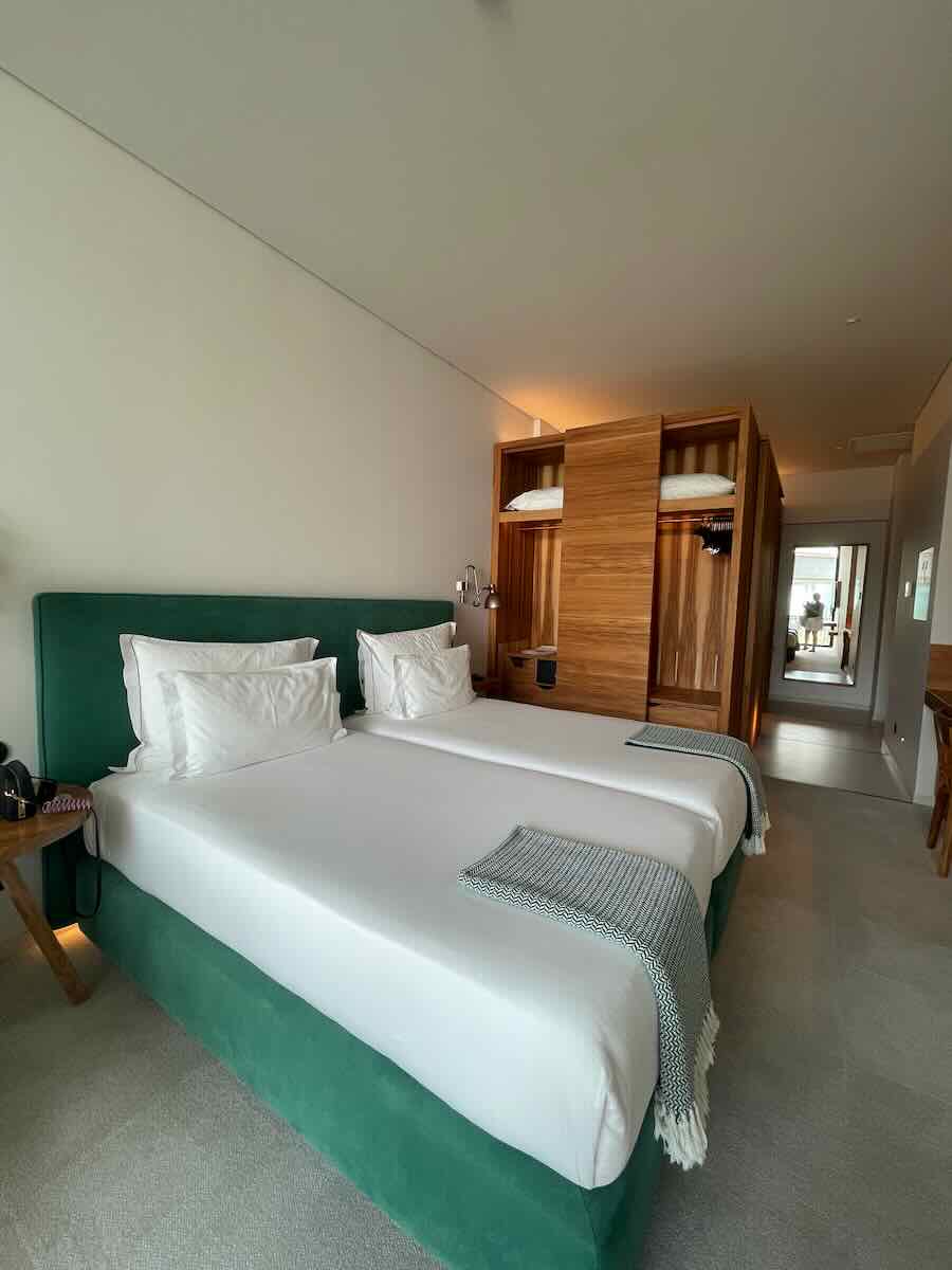 A modern hotel room in Corfu featuring twin beds with white linens, green upholstered bed frames, wooden wardrobe with sliding doors, and minimal decor.