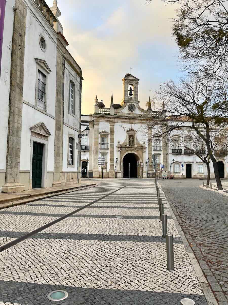 A picturesque square in Faro, Portugal, featuring a historic white church with a bell tower and clock at its center. The foreground showcases a patterned cobblestone pavement, flanked by elegant white buildings and bare trees under a soft, golden sky.