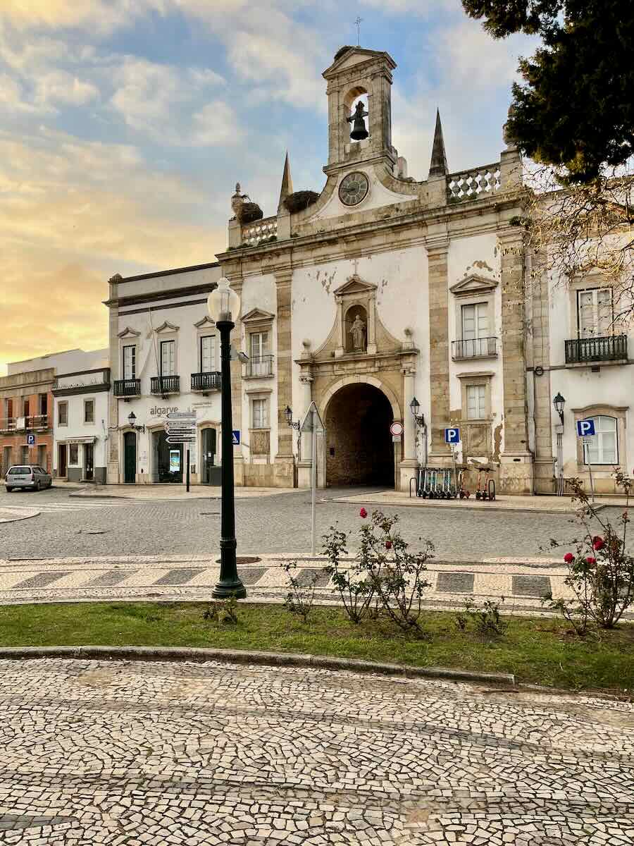 A historic archway in Faro, Portugal, adorned with a bell tower and a clock, surrounded by white buildings. The foreground features a cobblestone street, a lamp post, and a small rose garden, with the sky painted in soft golden hues of sunset.