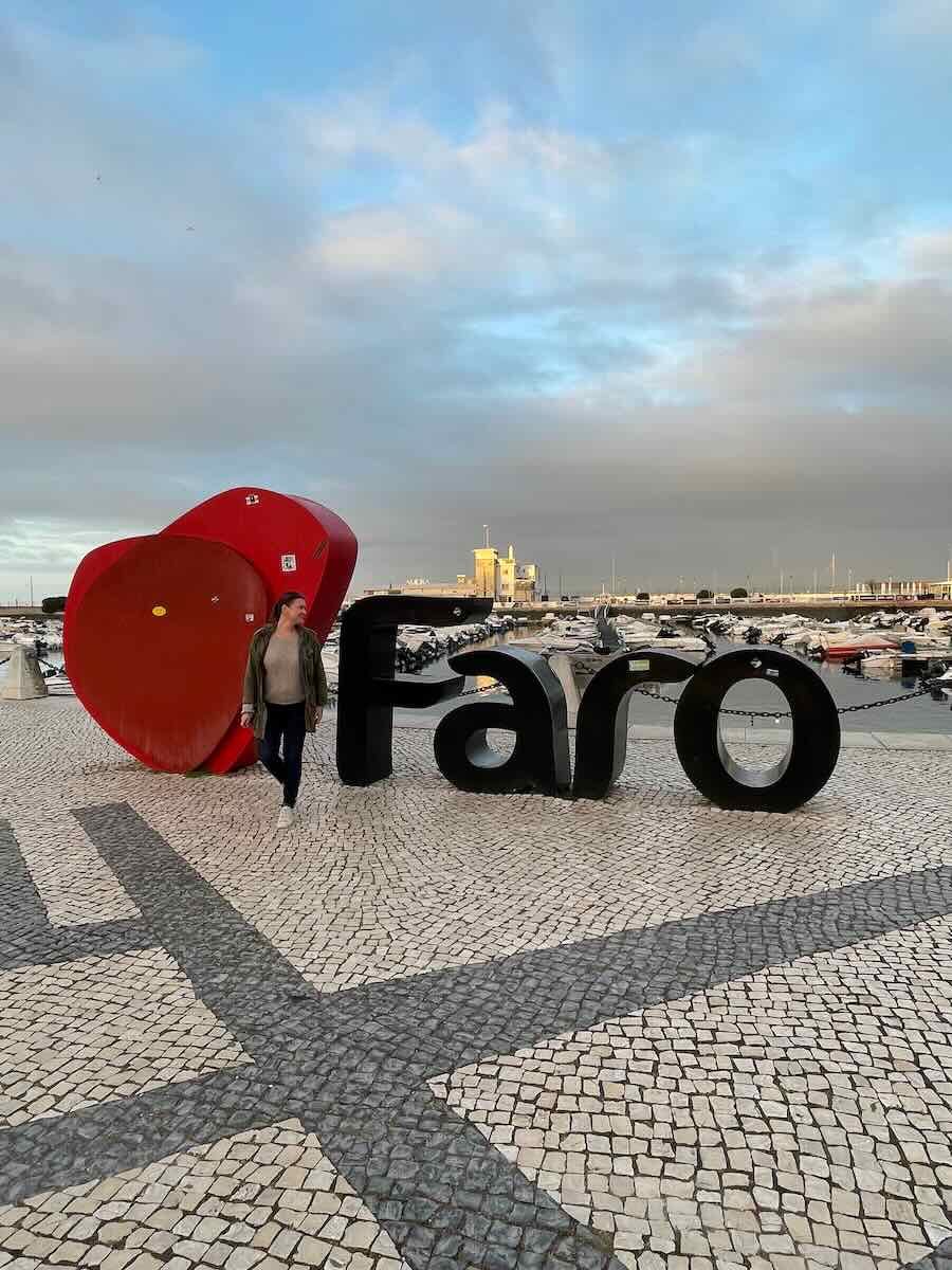 A modern "Faro" sign near the marina in Faro, Portugal, with a bold red heart symbolizing love for the city. The background features moored boats, a harbor, and a cloudy sky during sunset, adding warmth and vibrancy to the scene.