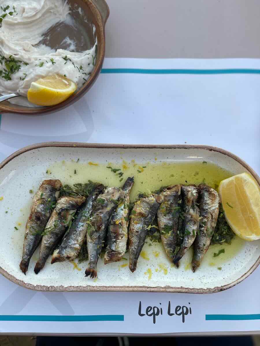 The image shows grilled sardines with herbs and olive oil, served with a lemon wedge and a side of creamy spread, likely tzatziki. The restaurant is named "Lepi Lepi."