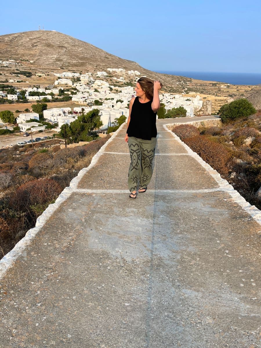 Melissa walks along a stone pathway on a Greek island, brushing her hair back as she gazes into the distance. She wears a black sleeveless top and green patterned pants, with the white-washed buildings of a traditional village spread across the hillside behind her. The dry, rugged landscape contrasts with the deep blue of the Aegean Sea on the horizon.