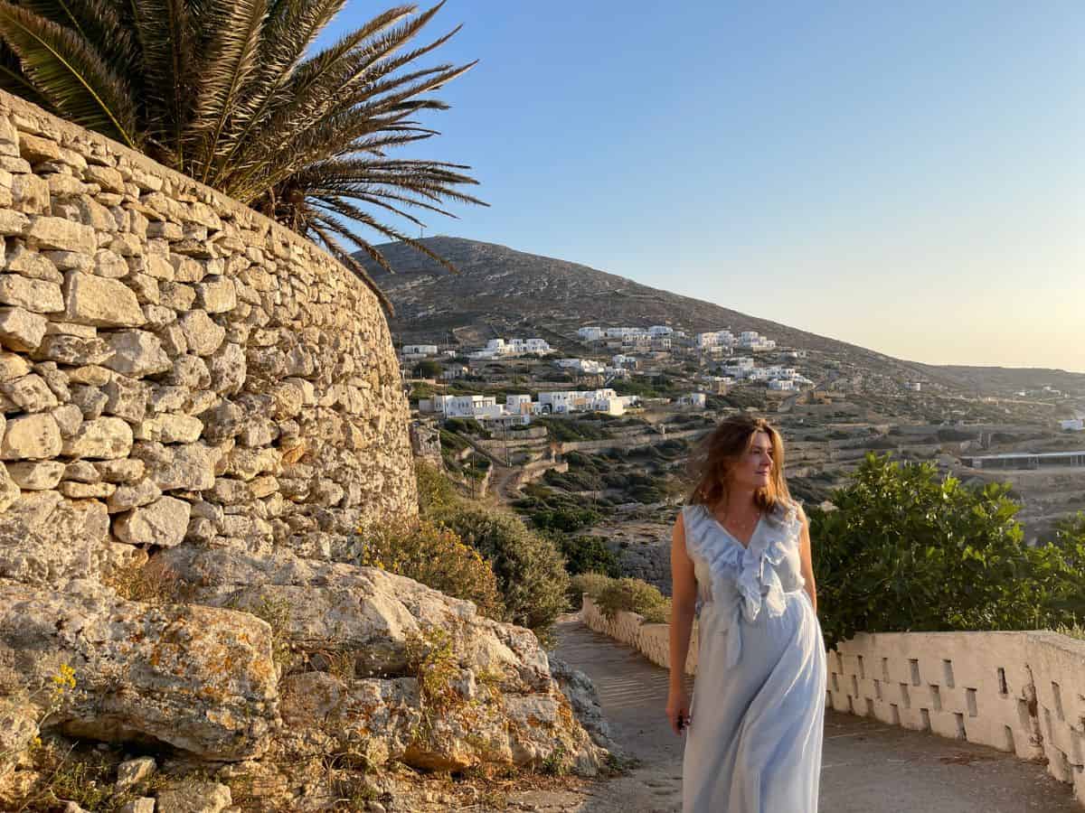 A woman in a flowing white dress stands on a hillside pathway near a traditional stone wall on Folegandros, an affordable Greek island close to Mykonos. The background features a typical Cycladic landscape with white houses dotting the arid hills during golden hour.