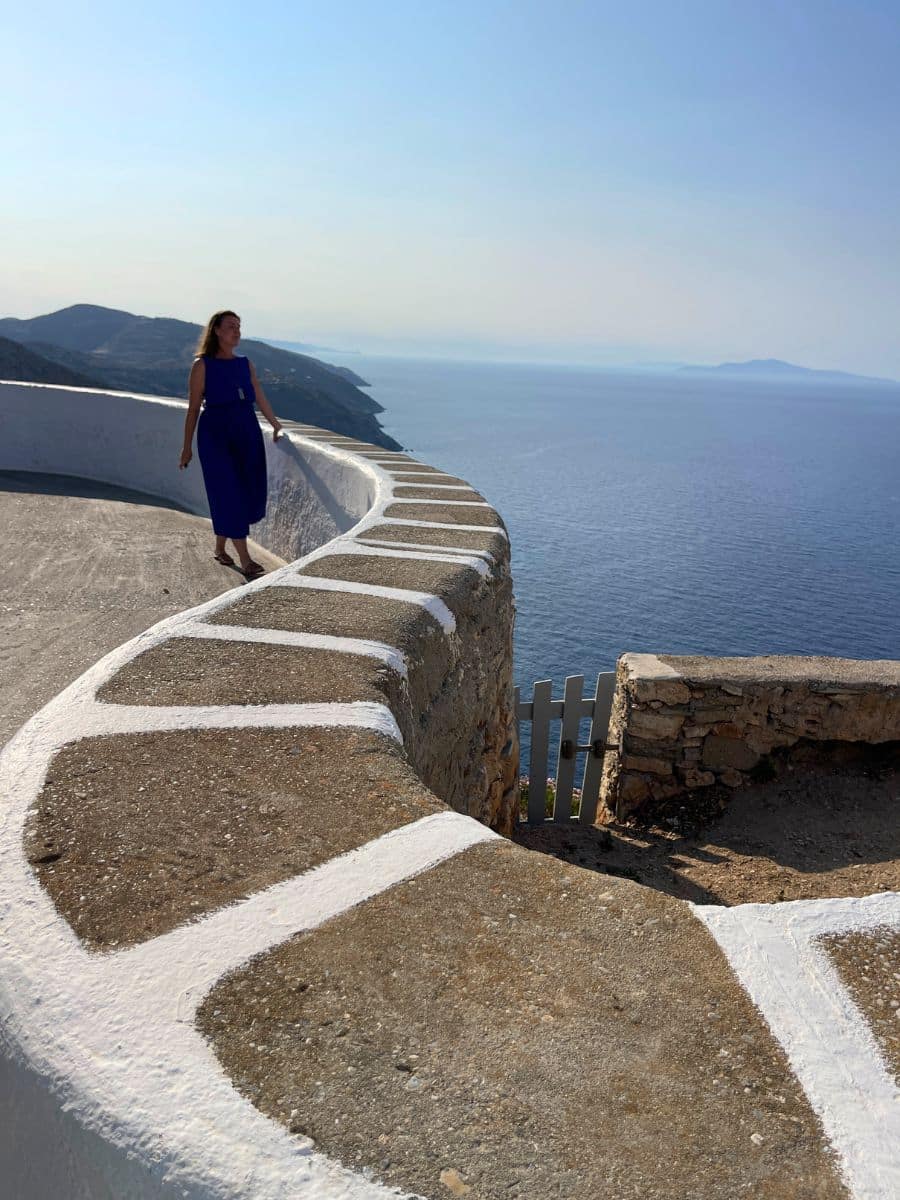 A woman in a flowing blue dress walks along a curved whitewashed stone wall, overlooking the deep blue Aegean Sea in Folegandros, Greece. The late afternoon sun casts a warm glow, highlighting the rugged coastline and distant islands in the background. The traditional Cycladic architecture blends seamlessly with the natural beauty of the island.