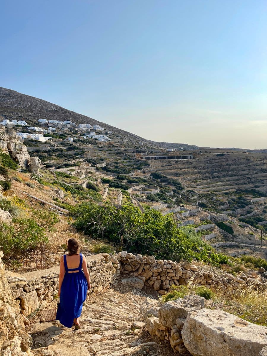 Melissa in a blue dress walks down a rugged stone path in Folegandros, Greece, with her back turned to the camera. The warm afternoon sunlight highlights the earthy tones of the rocky landscape, interspersed with patches of green vegetation. Traditional whitewashed houses are scattered across the hillside in the background, forming a picturesque Cycladic village. The winding terraces and dry-stone walls showcase the island’s characteristic charm and history.
