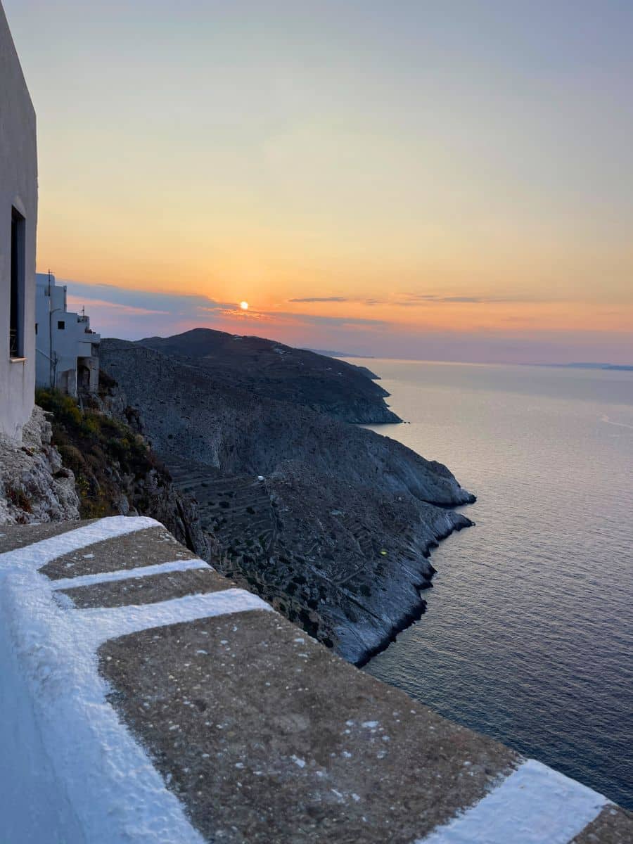 A breathtaking sunset view from a cliffside in Folegandros, Greece, with white-washed buildings on the left, rugged hills in the distance, and the calm Aegean Sea stretching to the horizon.