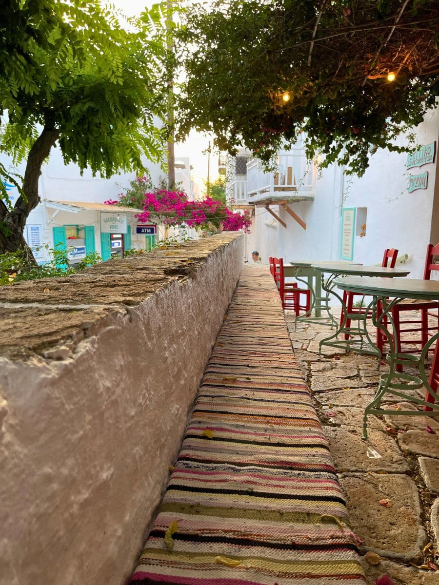 A cozy outdoor seating area in Folegandros, Greece, featuring a long cushioned bench with a striped rug, red and green chairs, and a charming white-washed backdrop adorned with bougainvillea and greenery under warm string lights.