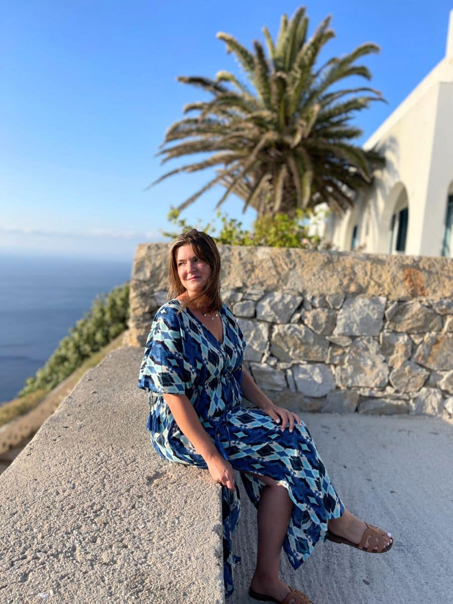 Melissa in a flowing blue patterned dress sits on a stone ledge in Folegandros, Greece, enjoying the coastal view. A tall palm tree and a white Mediterranean-style building with arched windows are behind her, while the deep blue sea stretches into the horizon. The golden sunlight enhances the serene and picturesque setting.
