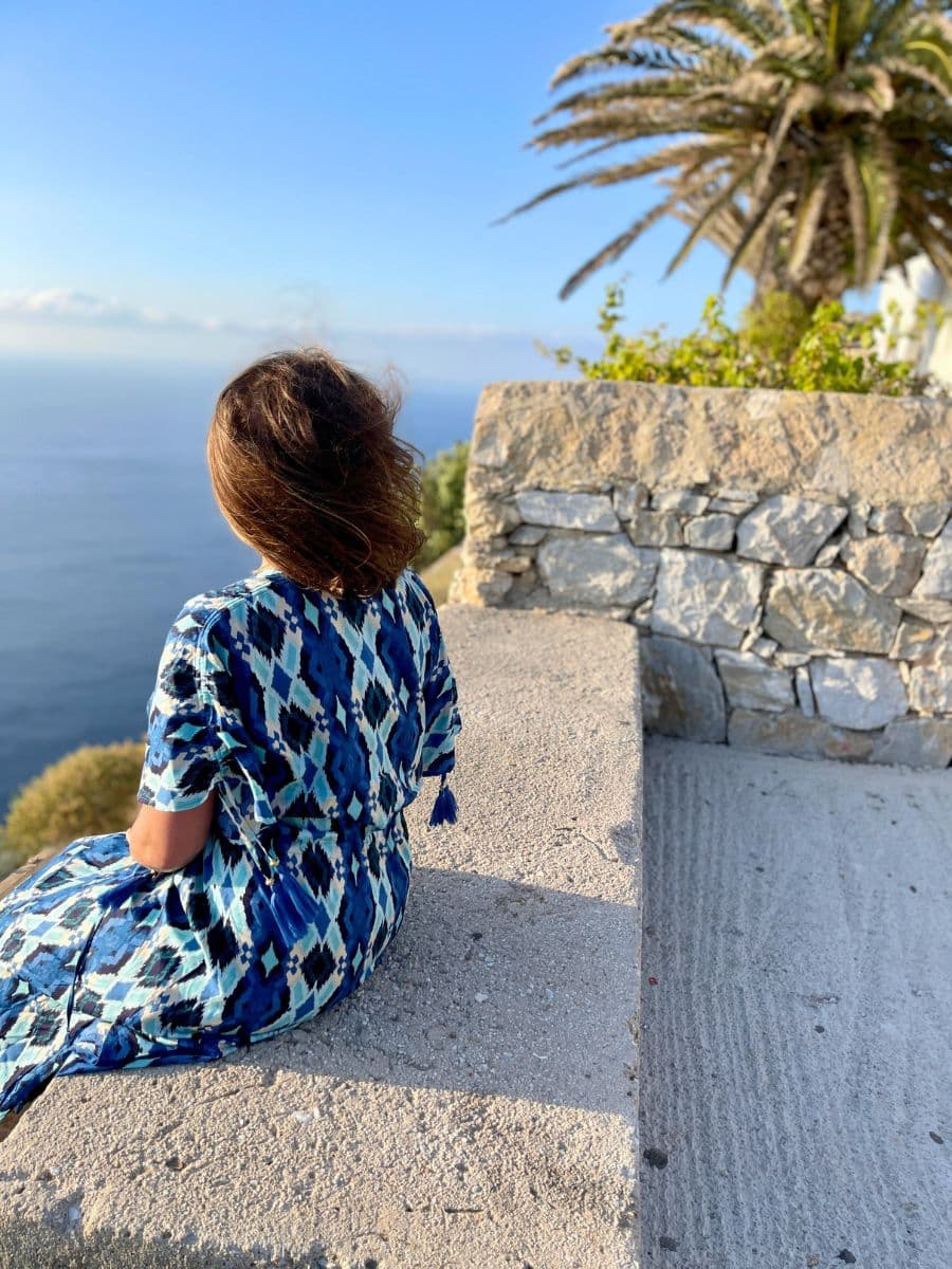 Melissa in a flowing blue patterned dress sits on a stone ledge, gazing out at the vast, deep blue sea in Folegandros, Greece. Her hair is gently tousled by the breeze, and the golden sunlight highlights the peaceful and contemplative moment. A palm tree and a stone wall add to the serene Mediterranean atmosphere.