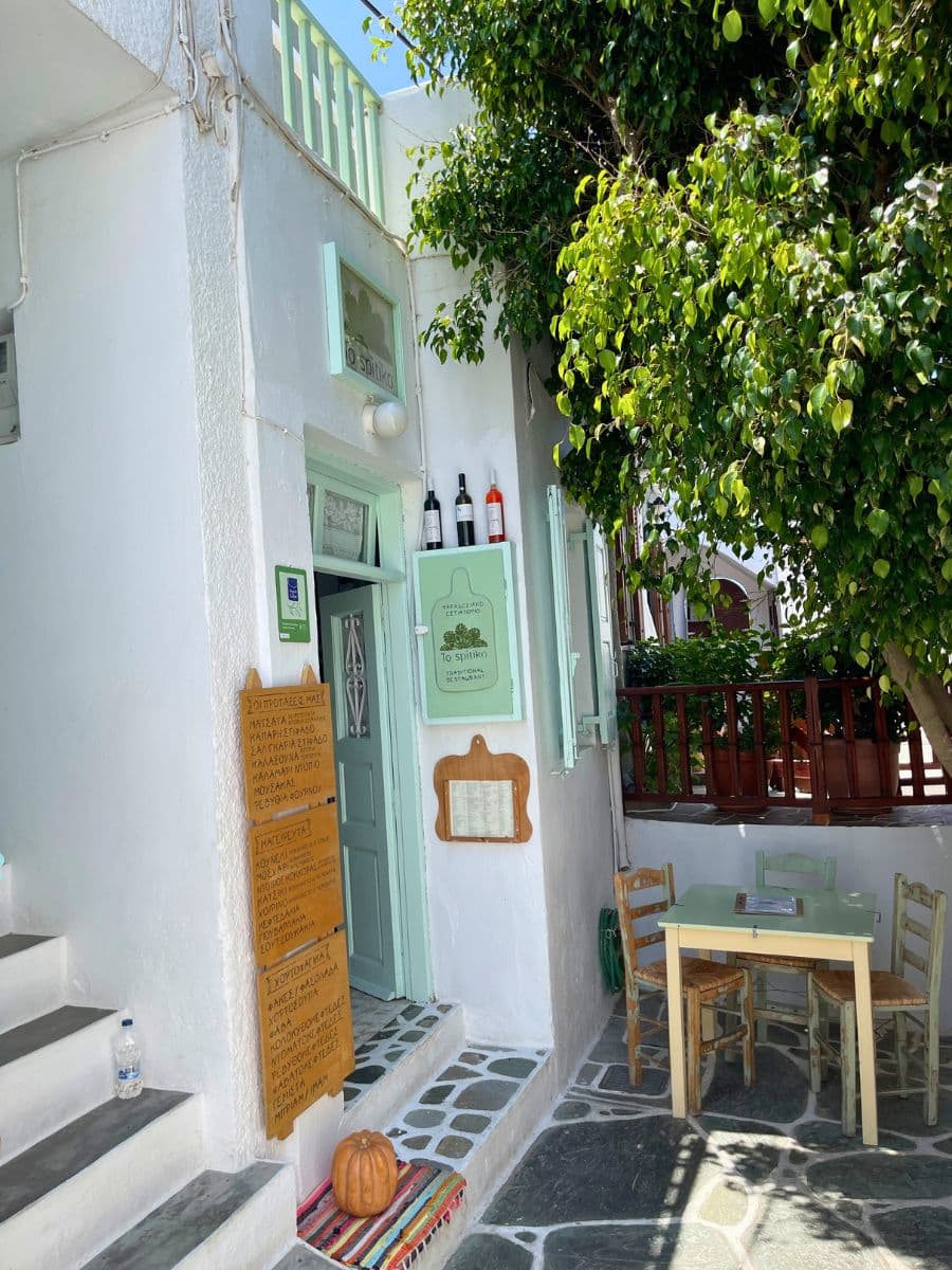 A quaint traditional restaurant in Folegandros with pastel green doors and signage, wooden menu boards listing Greek dishes, a small outdoor seating area with rustic chairs, and a tree providing shade.