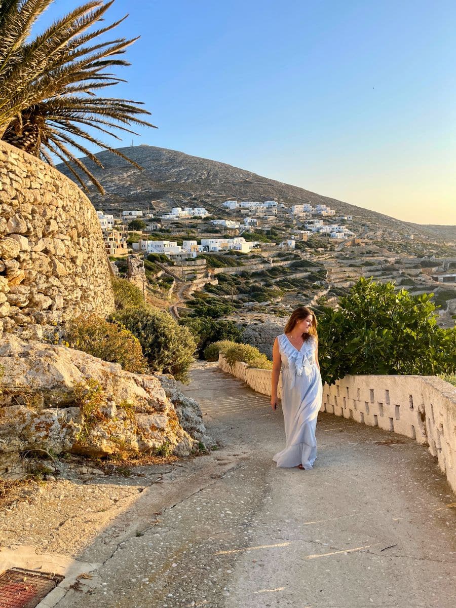 Melissa in a flowing light blue dress walks along a sunlit path in Folegandros, Greece. The golden hour light bathes the rugged landscape, with white-washed houses scattered across the hillside in the background.