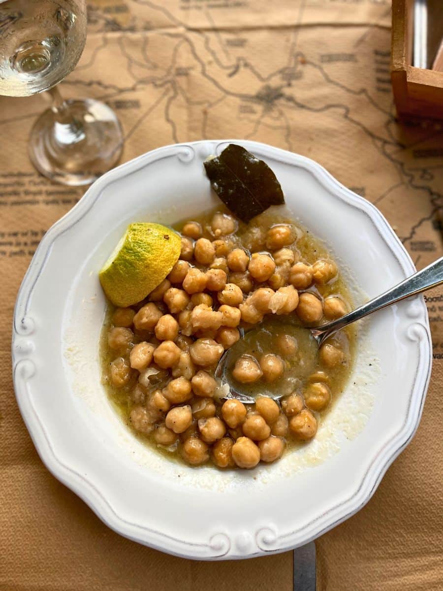 A plate of traditional Greek revithada, a slow-cooked chickpea stew, served in a white dish with a bay leaf and a wedge of lemon on the side. A spoon rests in the bowl, and a glass of white wine sits on the table, which is covered with a paper map-themed placemat.