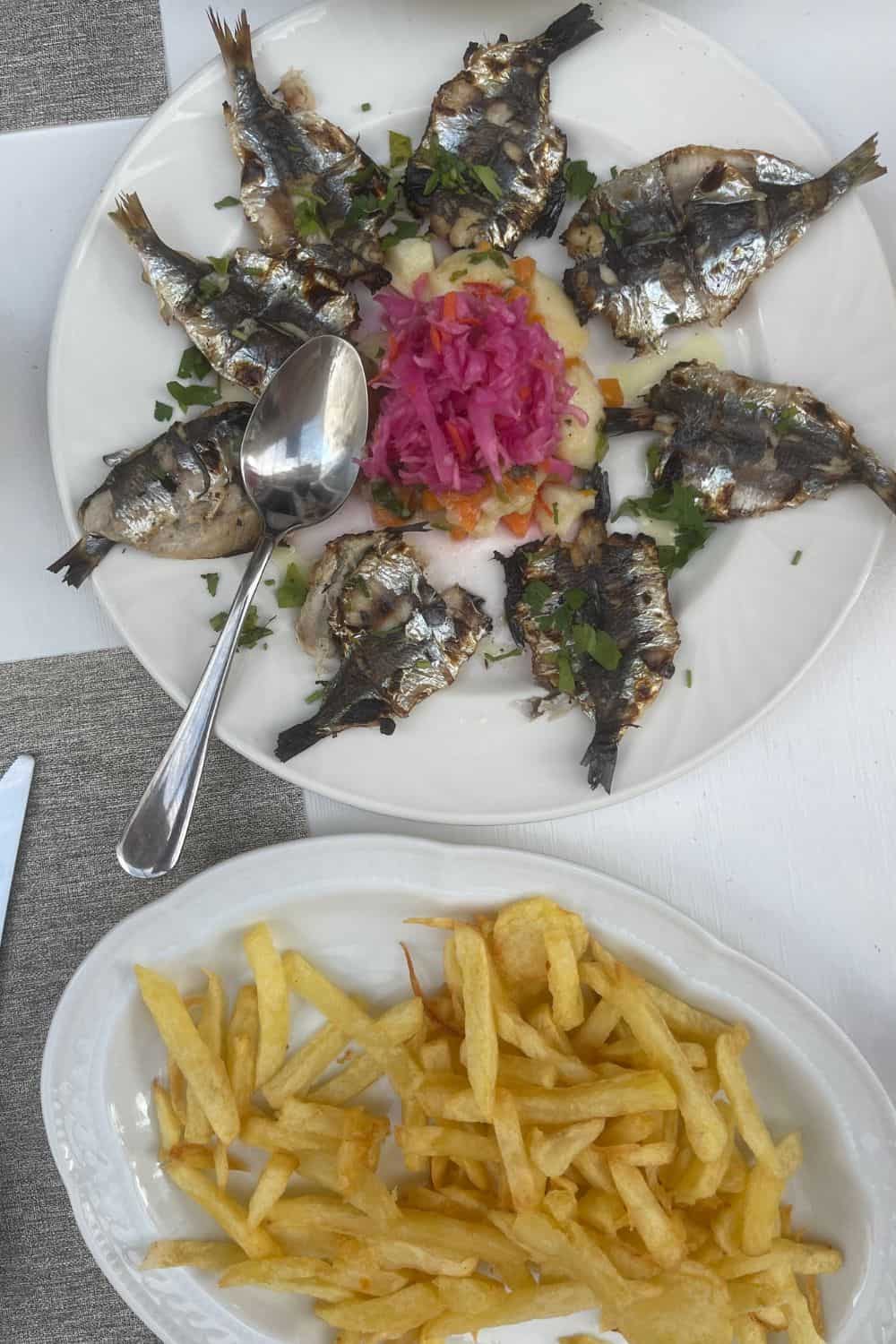 An elegant dining setting with Mediterranean cuisine served in a restaurant in Syros, Greece. The table features a hearty slice of grilled squash on a bed of creamy sauce, and a smooth potato purée topped with a succulent tomato. To the side, a tray with rustic bread and olive oil dip, complementing the dishes. The fine dining experience is accentuated by the neat placement of cutlery on a gray napkin and the simplicity of the glassware, inviting guests to a culinary journey.