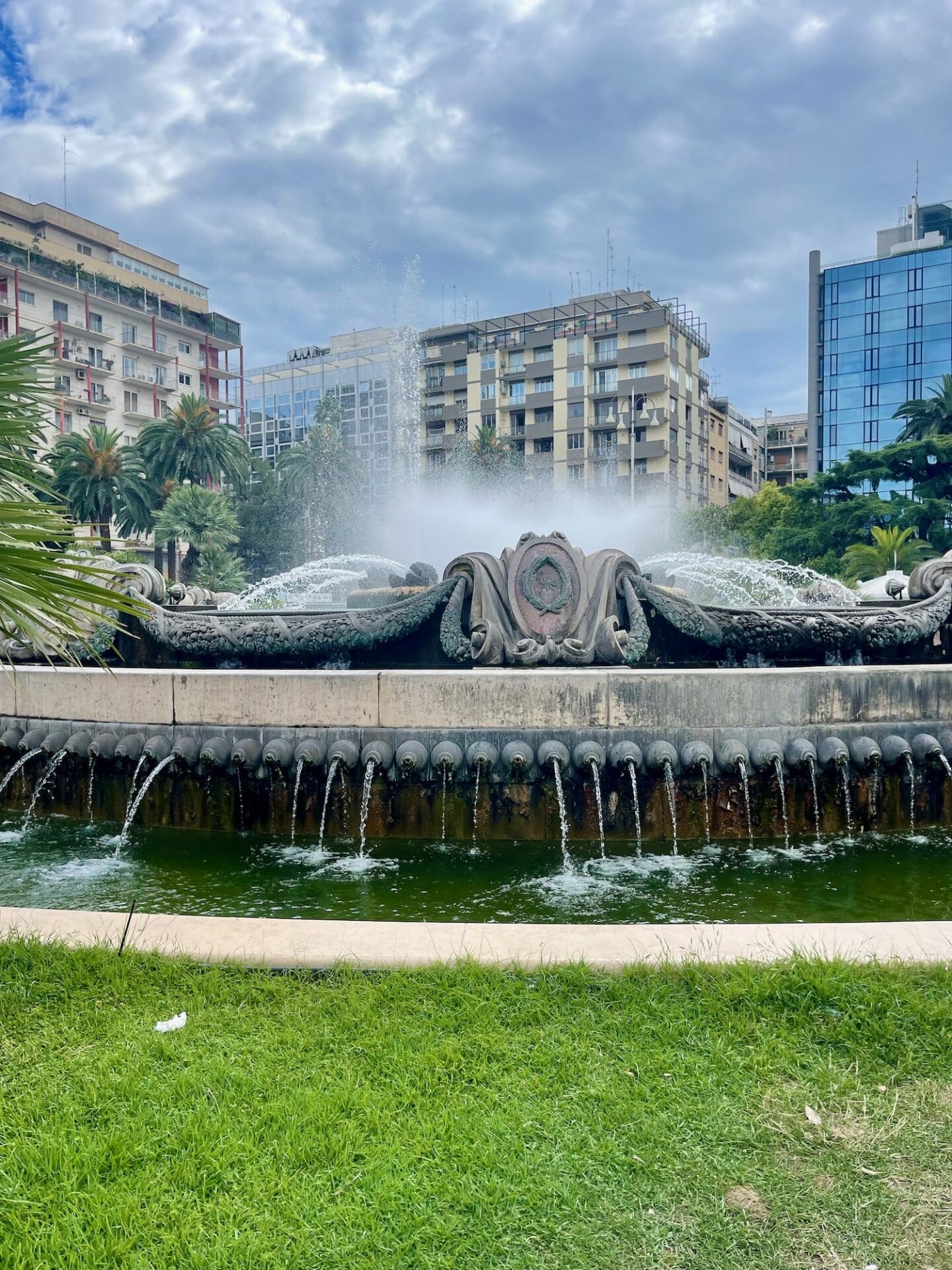 A decorative fountain in Bari, Italy, with water jets spraying into the air. The fountain is surrounded by lush green grass and set against a backdrop of tall buildings and palm trees under a cloudy sky.