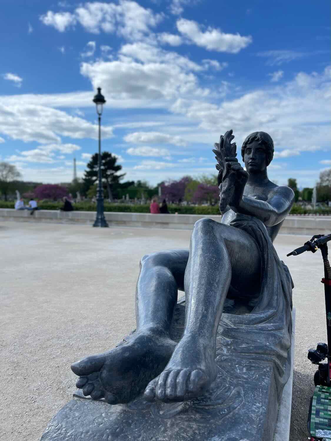 A close-up of a bronze statue of a reclining woman in the Jardin des Tuileries in Paris, with a bright blue sky and fluffy white clouds in the background.