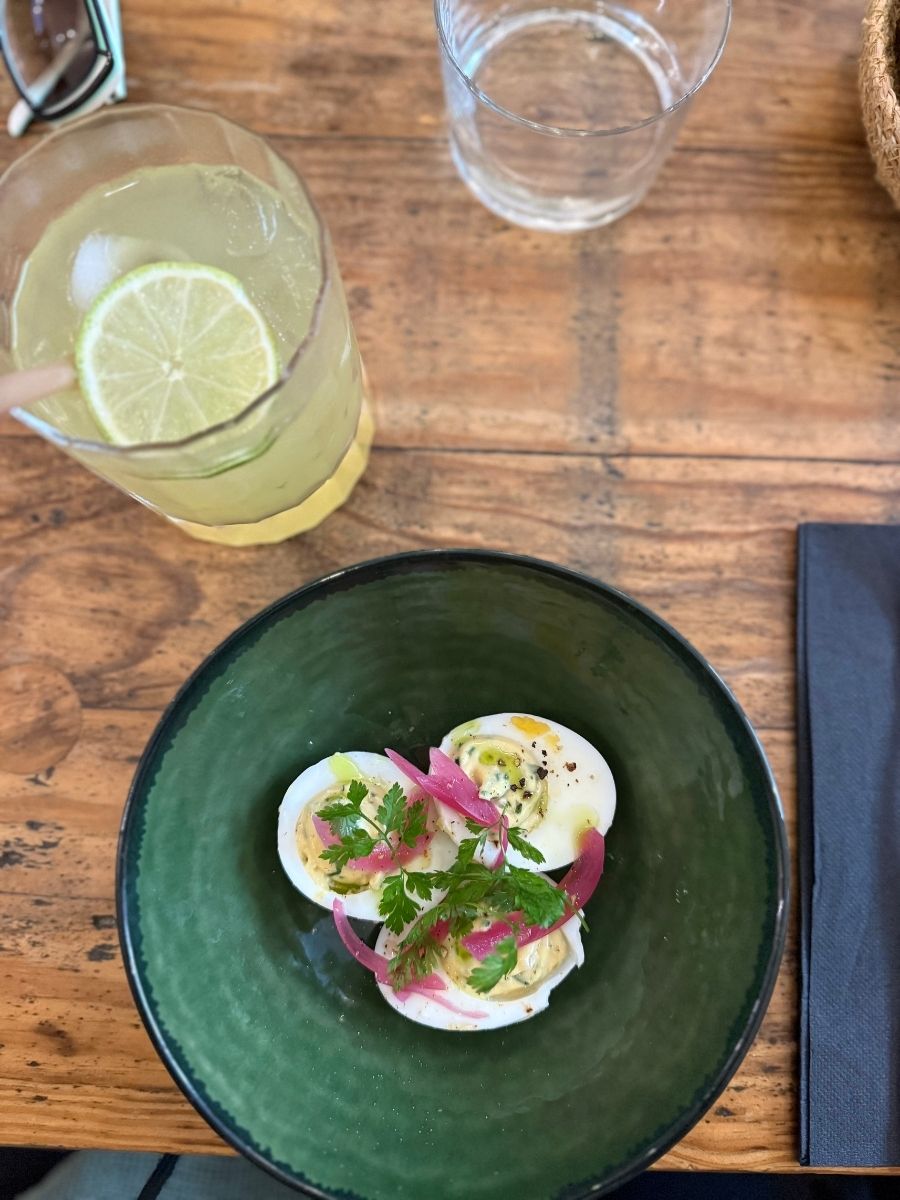 Close-up of a green ceramic bowl with deviled eggs topped with herbs and pickled onions, served with a refreshing lime drink at a rustic wooden table in a Bordeaux café.