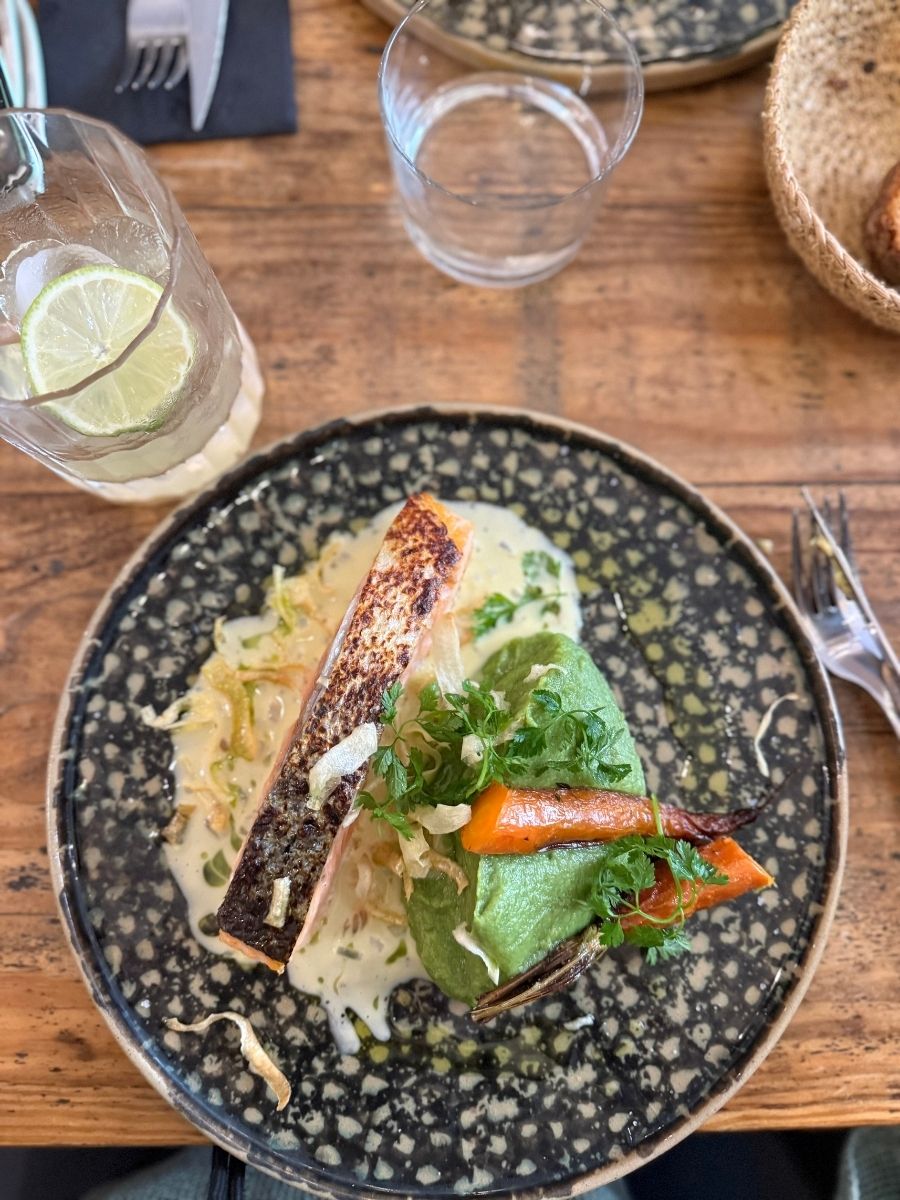 Beautifully plated salmon dish with creamy leeks, greens, and grilled vegetables at a cozy café in Bordeaux, accompanied by a lime drink and rustic wooden table setting.