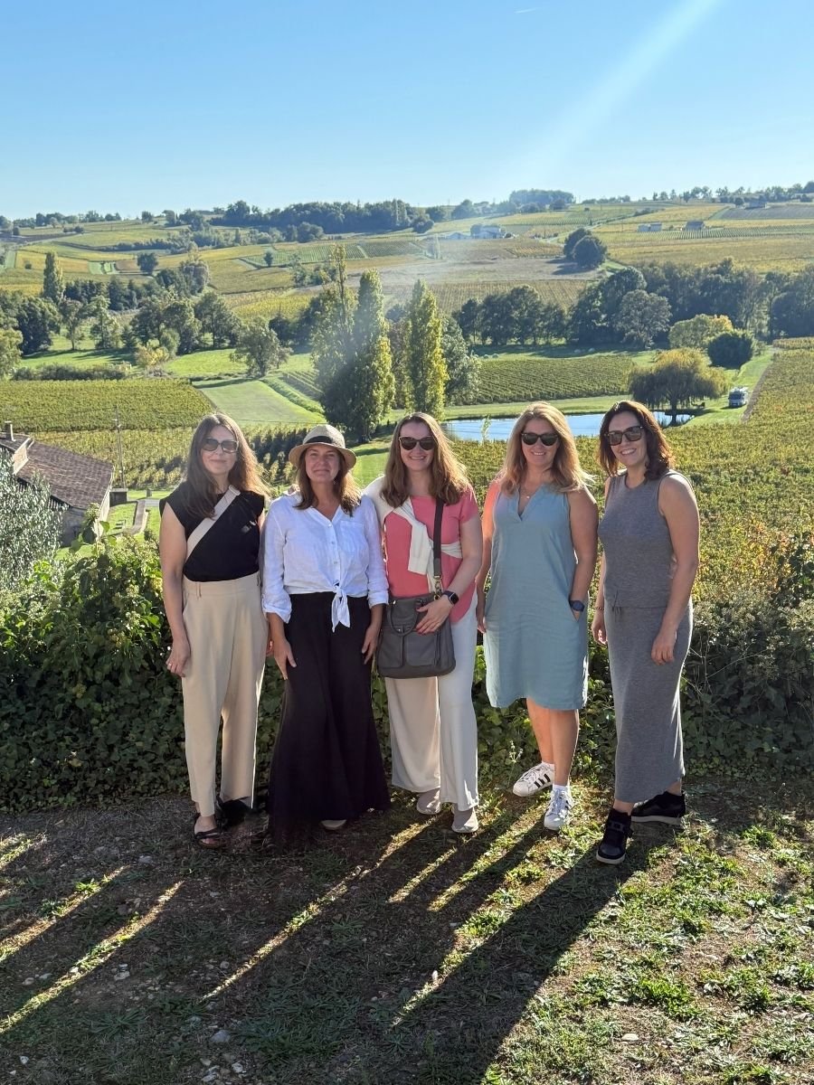 A group of five women posing together in front of the picturesque vineyards of Saint-Émilion, Bordeaux, France, under the clear afternoon sun.
