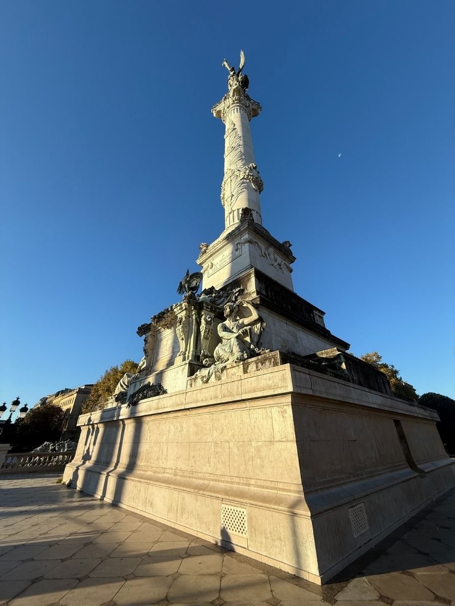 A low-angle view of the Monument aux Girondins in Bordeaux, France, with sculpted statues at the base and a clear blue sky in the background.