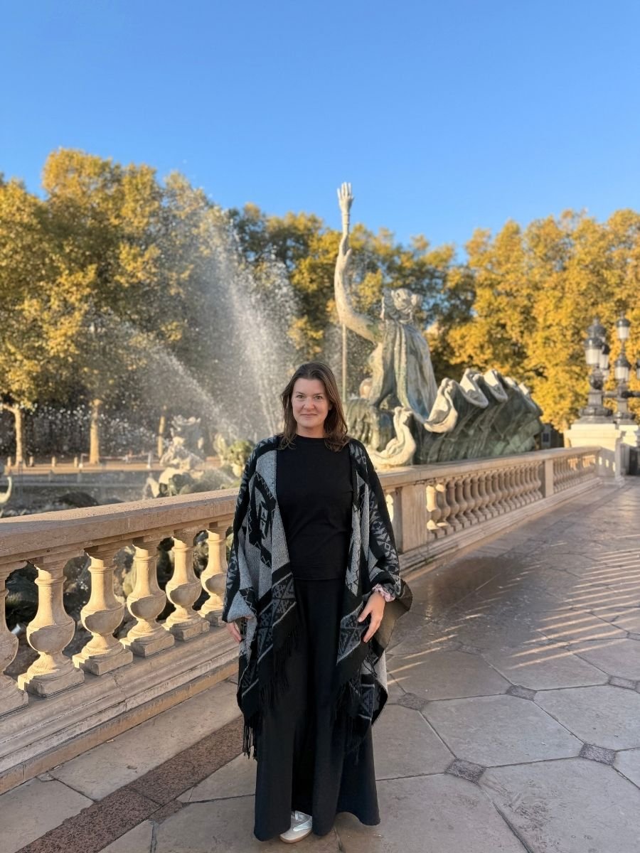 Melissa standing in front of a dramatic fountain at Place des Quinconces in Bordeaux, wearing a black outfit and patterned shawl, with golden autumn trees behind.