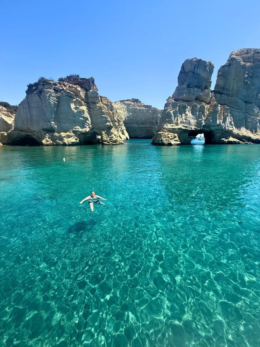 A swimmer floats peacefully in the unbelievably clear turquoise waters of Milos, surrounded by rocky coastal cliffs.