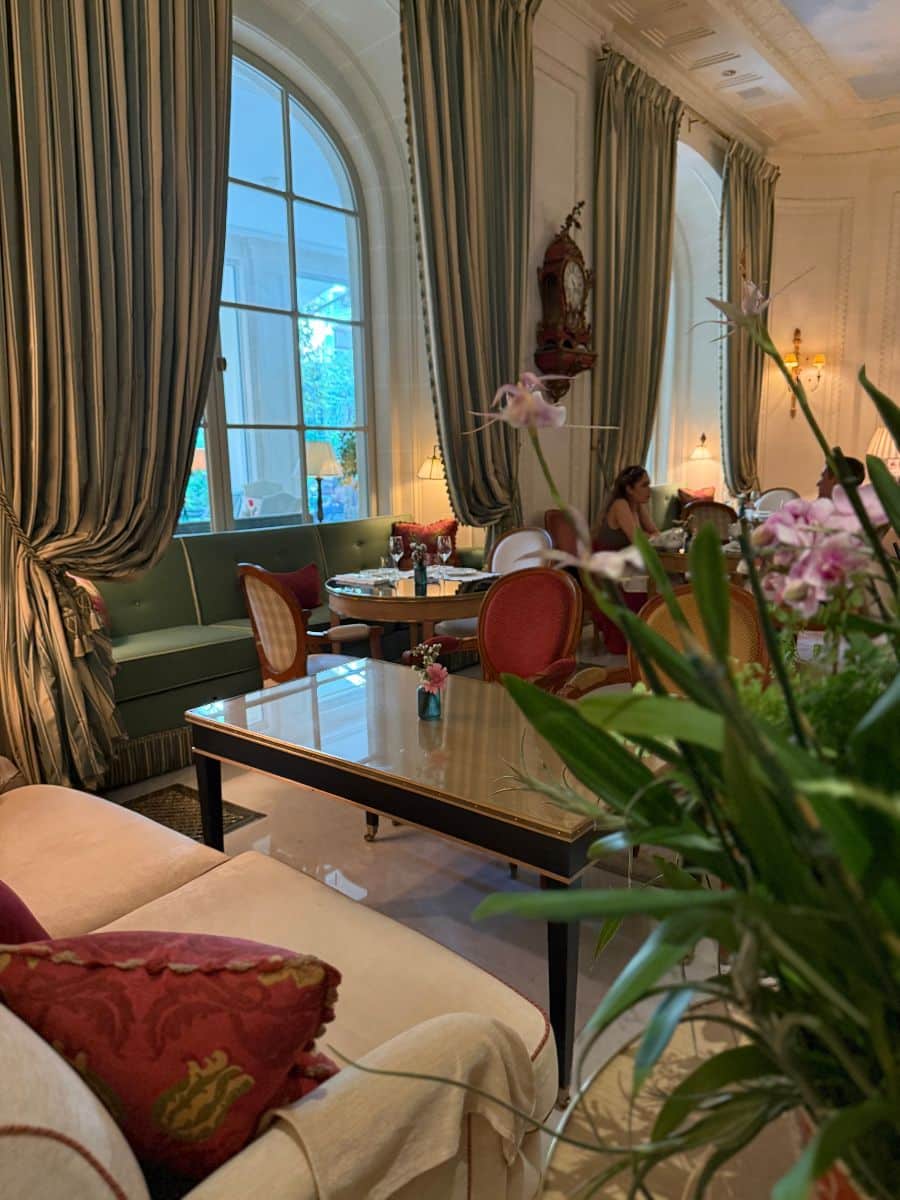 Elegant lounge with green drapes, floral arrangements, vintage clocks, and intimate seating in a Parisian hotel.