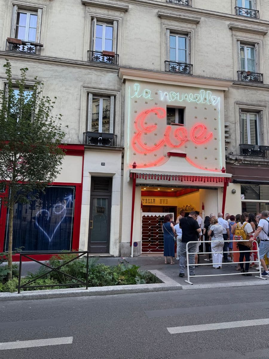 A group of people lined up outside the La Nouvelle Eve cabaret in Paris, under a bright neon sign on a classic Parisian building.