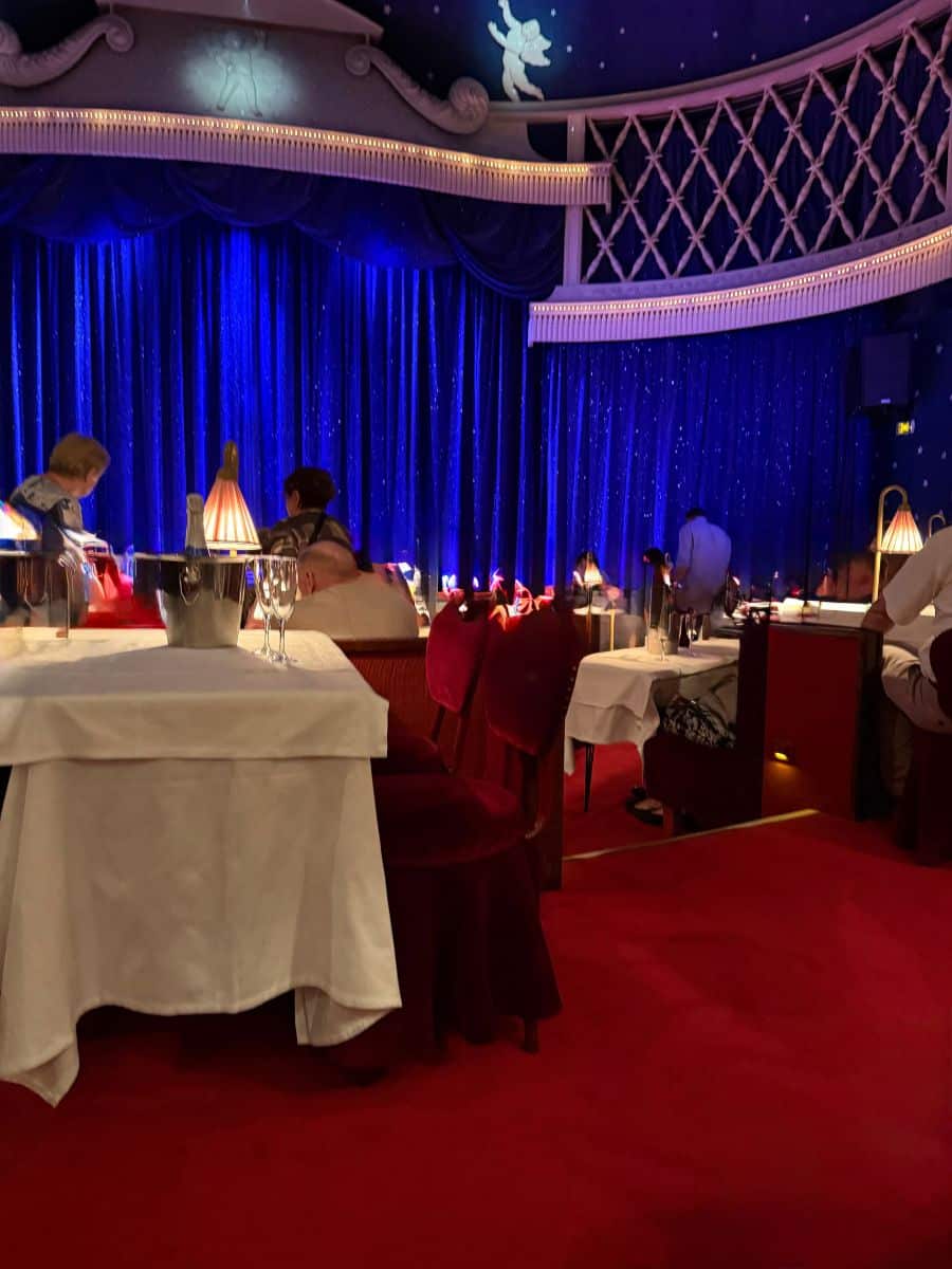 Elegant dining tables with white tablecloths and red velvet chairs set before a blue curtain stage at La Nouvelle Eve.