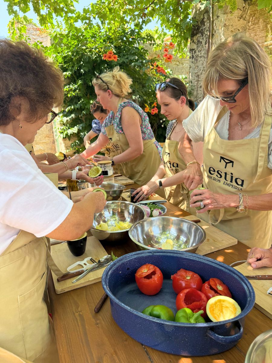 Women gathered around a wooden table in Naxos, Greece, preparing vegetables and traditional ingredients during an outdoor cooking class under a leafy pergola.