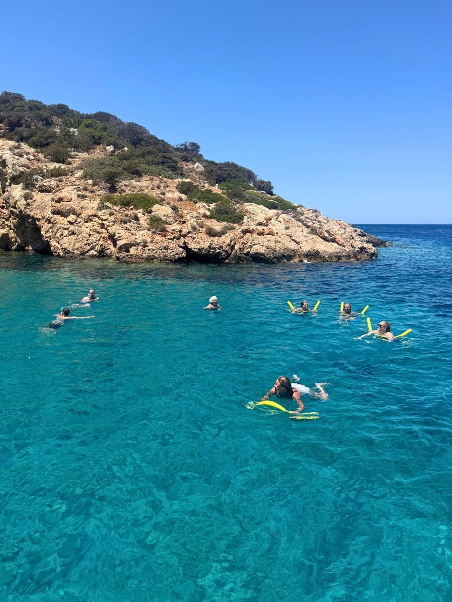 Group of women swimming and relaxing with pool noodles in crystal-clear turquoise waters near a rocky coastline in Naxos, Greece.