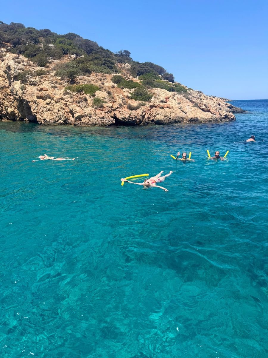 Travelers floating with pool noodles in the crystal-clear turquoise waters off the rocky coast of Naxos, Greece, under a bright blue sky.