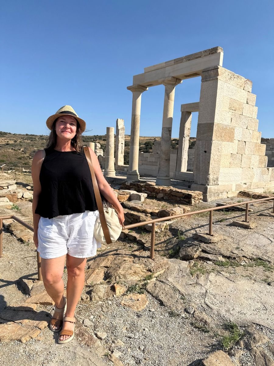 Melissa in a straw hat and summer outfit stands in front of the ancient marble Temple of Demeter ruins in Naxos, Greece, on a sunny day.