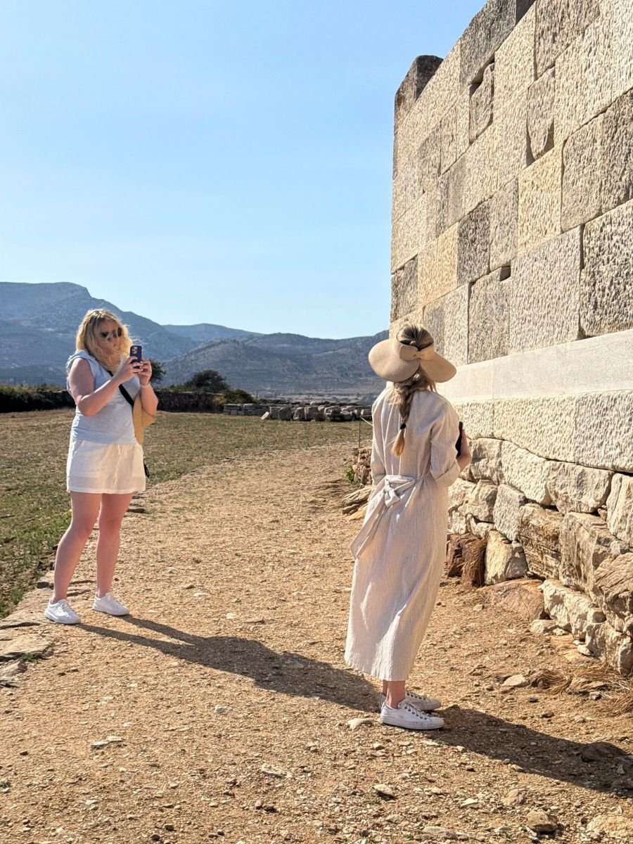Two women exploring ancient stone ruins in Naxos, Greece—one posing near a wall in a sunhat and dress, the other capturing the moment with her phone under clear blue skies.