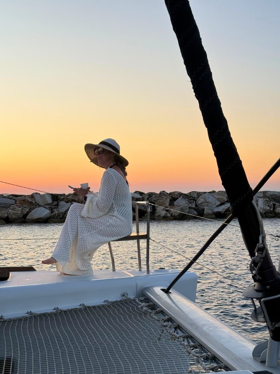 Woman relaxing on a catamaran at sunset in Naxos, Greece, wearing a sunhat and crochet cover-up, holding a cup and phone while enjoying the serene sea view.