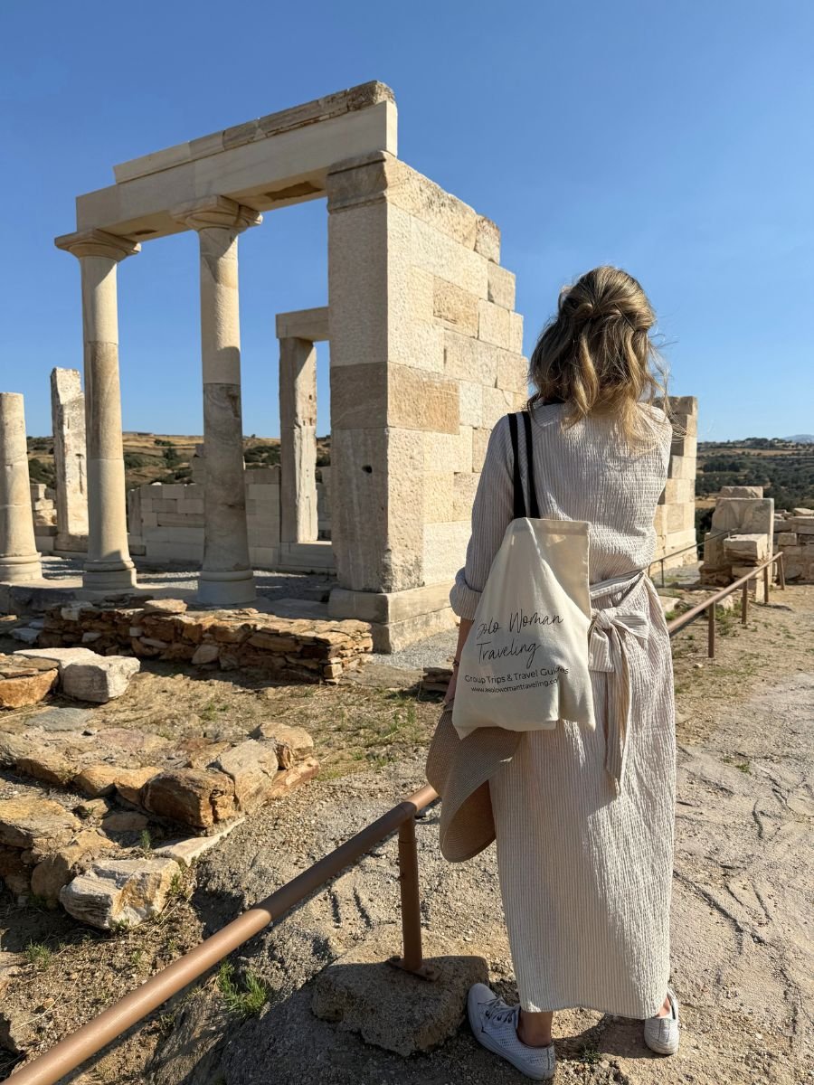 A woman in a striped dress and sneakers stands with her back to the camera, holding a sunhat and wearing a tote bag that reads "Solo Woman Traveling" while admiring the ancient Temple of Demeter ruins in Naxos, Greece, under a clear blue sky.