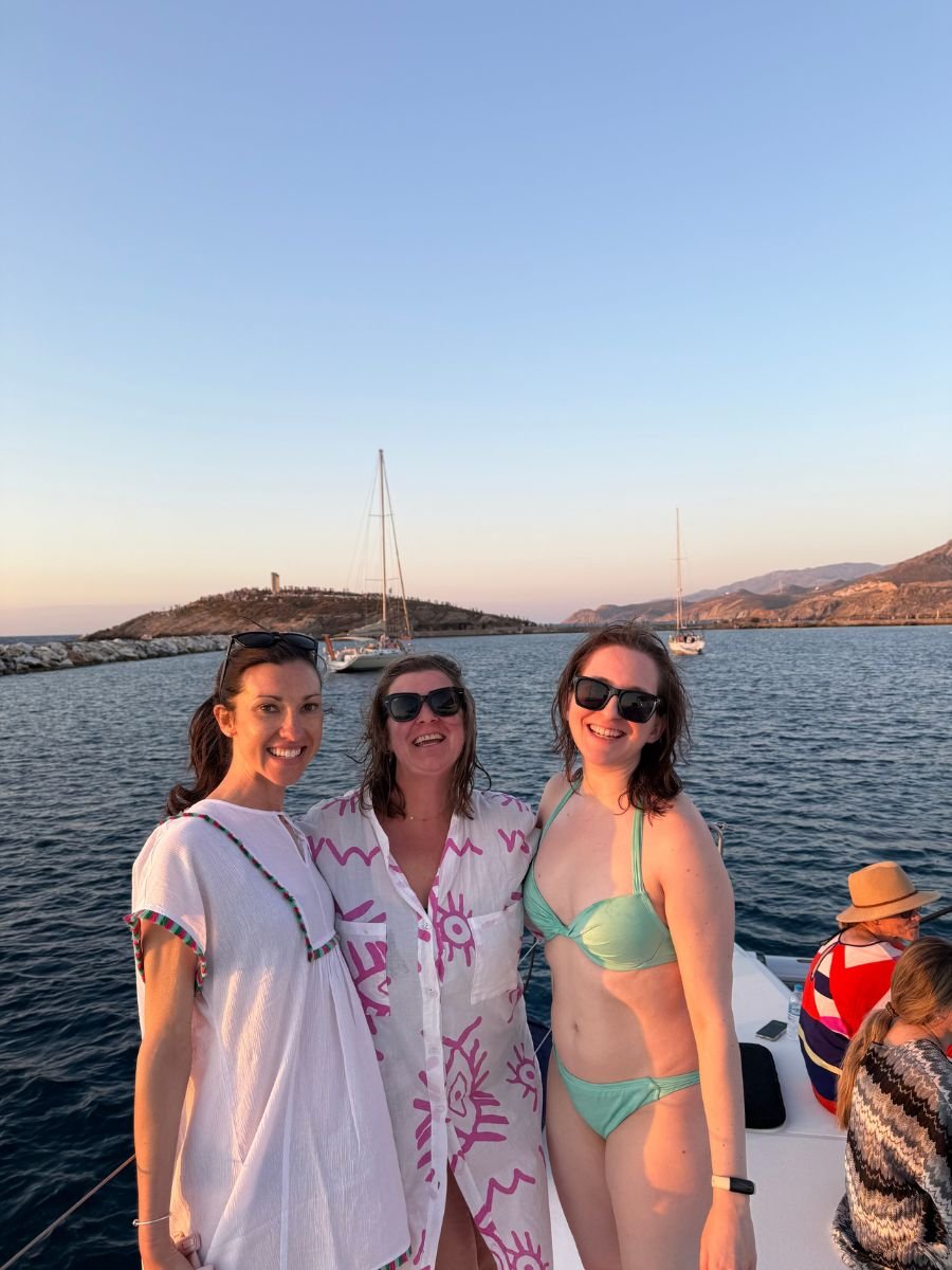 Three smiling women pose together on a boat during sunset in Naxos, Greece. One wears a white beach dress, another a pink-patterned cover-up, and the third a mint green bikini. The calm sea and distant sailboats are visible in the background.