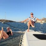 Two women relax on the deck of a catamaran in Naxos, Greece. One lounges on the netting while holding her phone, and the other sits confidently on the bow with arms outstretched, enjoying the sun and sea breeze. Sailboats and rocky hills dot the background.