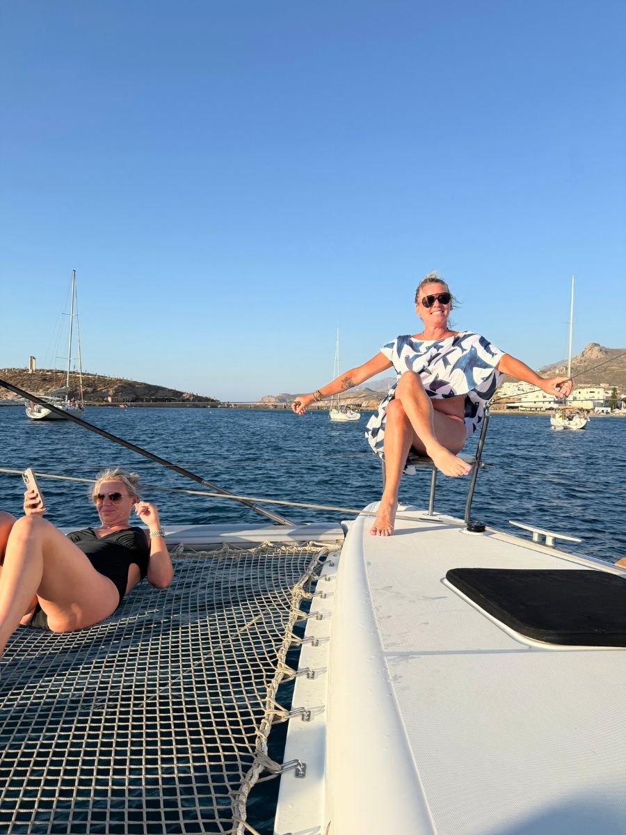 Two women relax on the deck of a catamaran in Naxos, Greece. One lounges on the netting while holding her phone, and the other sits confidently on the bow with arms outstretched, enjoying the sun and sea breeze. Sailboats and rocky hills dot the background.