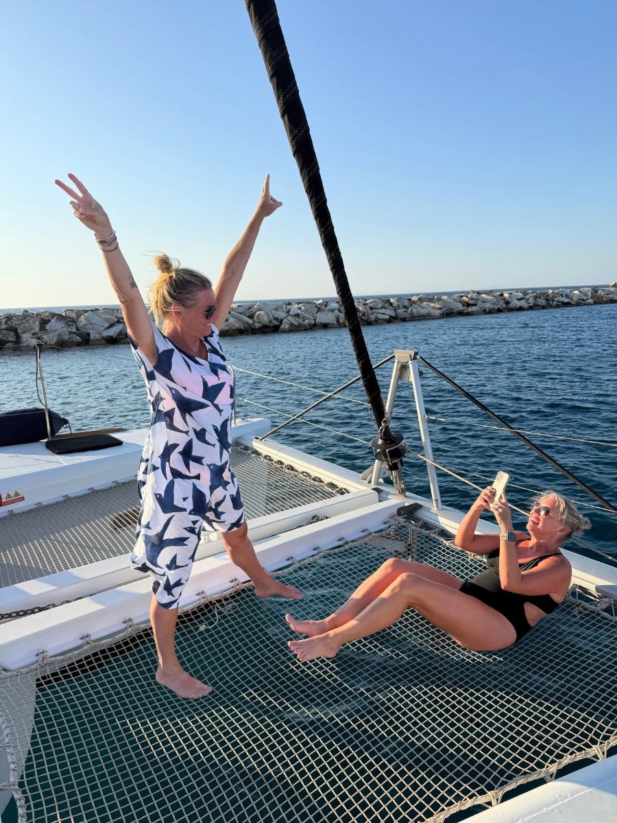 Two women enjoy playful moments on the netted deck of a catamaran in Naxos, Greece. One woman poses with arms raised in a victory sign, while the other, sitting in a black swimsuit, captures the moment on her phone. The sea and stone breakwater stretch out behind them under a clear blue sky.