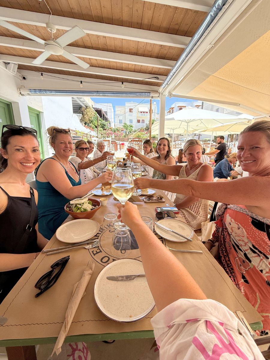 A joyful group of women raise their glasses in a celebratory toast during an outdoor lunch at a charming Greek taverna. The table is set with plates, fresh salads, and drinks, while the sunlit backdrop features white-washed buildings and blooming flowers, capturing the relaxed atmosphere of Naxos, Greece.