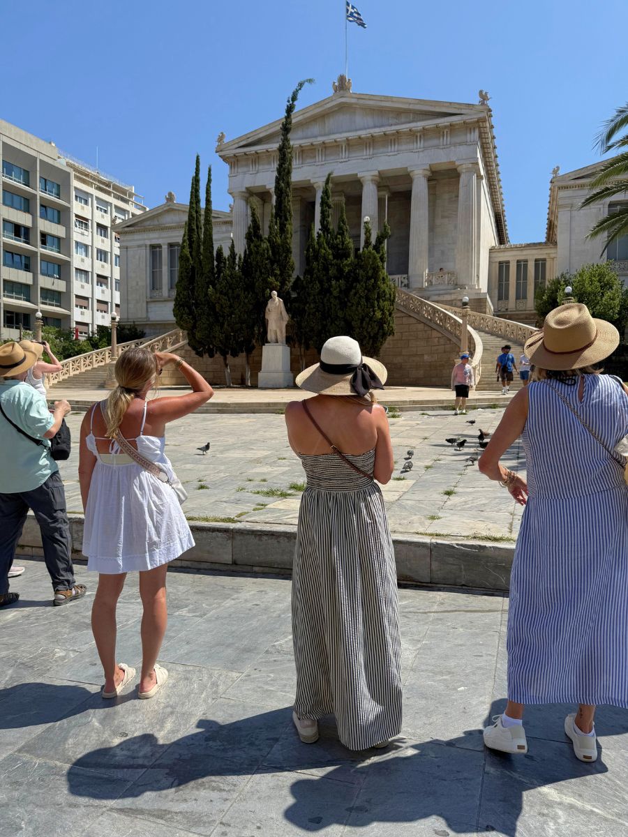 A group of tourists admire the grand neoclassical architecture of the National Library of Greece in Athens, with the Greek flag flying atop and tall cypress trees framing the marble staircase and statue.